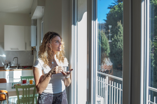 Woman standing in kitchen drinking a coffee and looking out the window